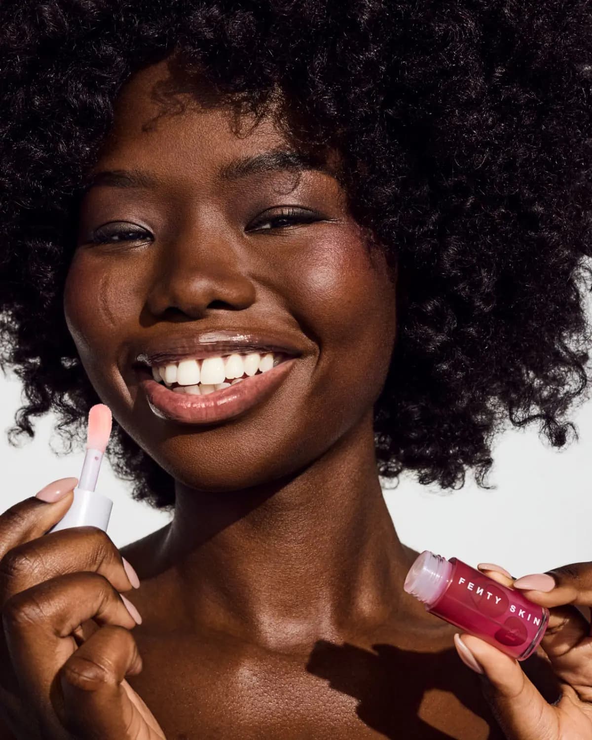 Smiling model with curly hair applying pink Fenty Skin lip product with wand