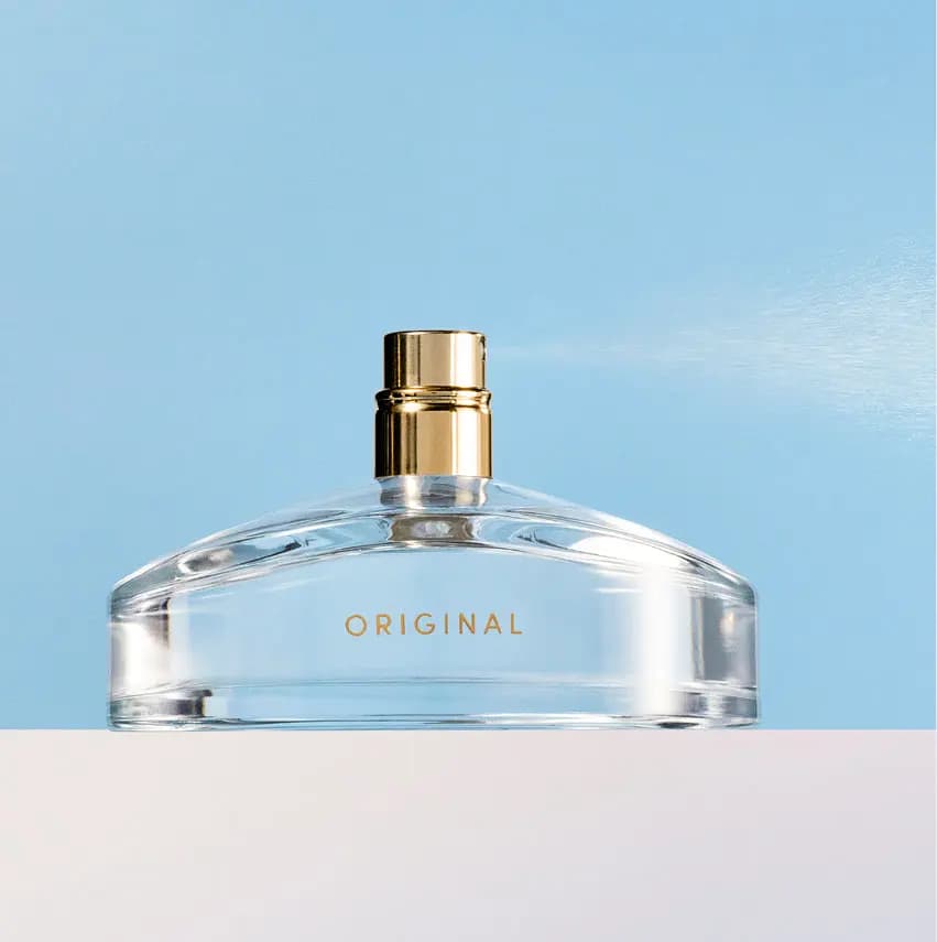 Uncapped clear bottle with gold spray nozzle misting fragrance against a sky-blue backdrop on a white ledge
