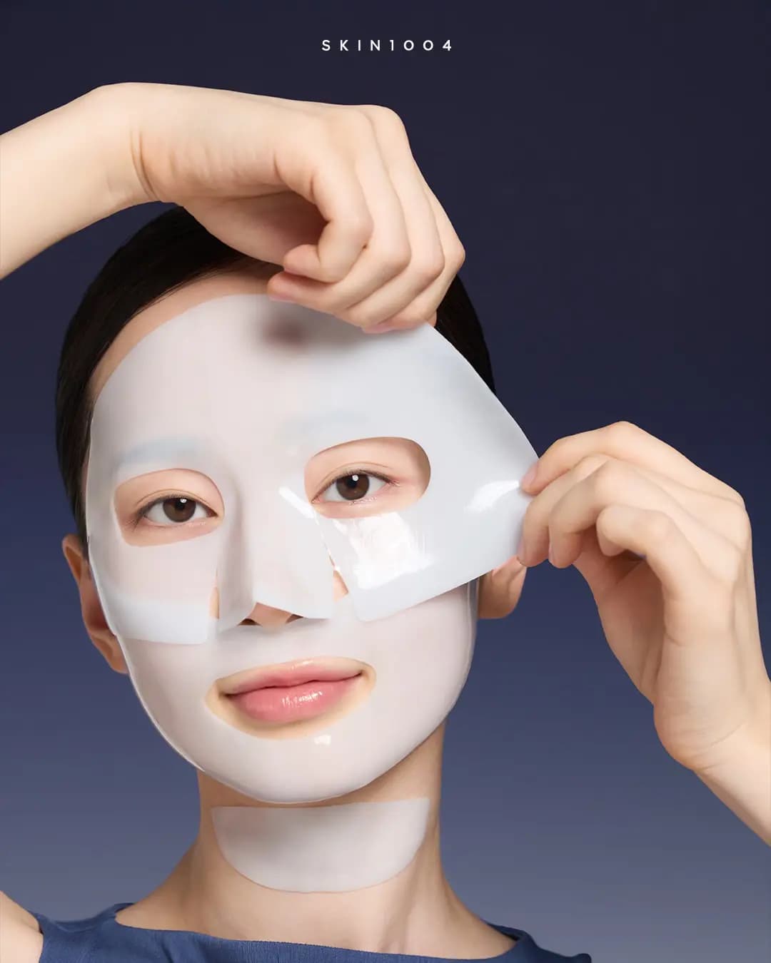 Model with dark hair lifting the translucent white SKIN1004 Hyalu-Teca gel mask off the face, with a matching strip on the neck, navy backdrop
