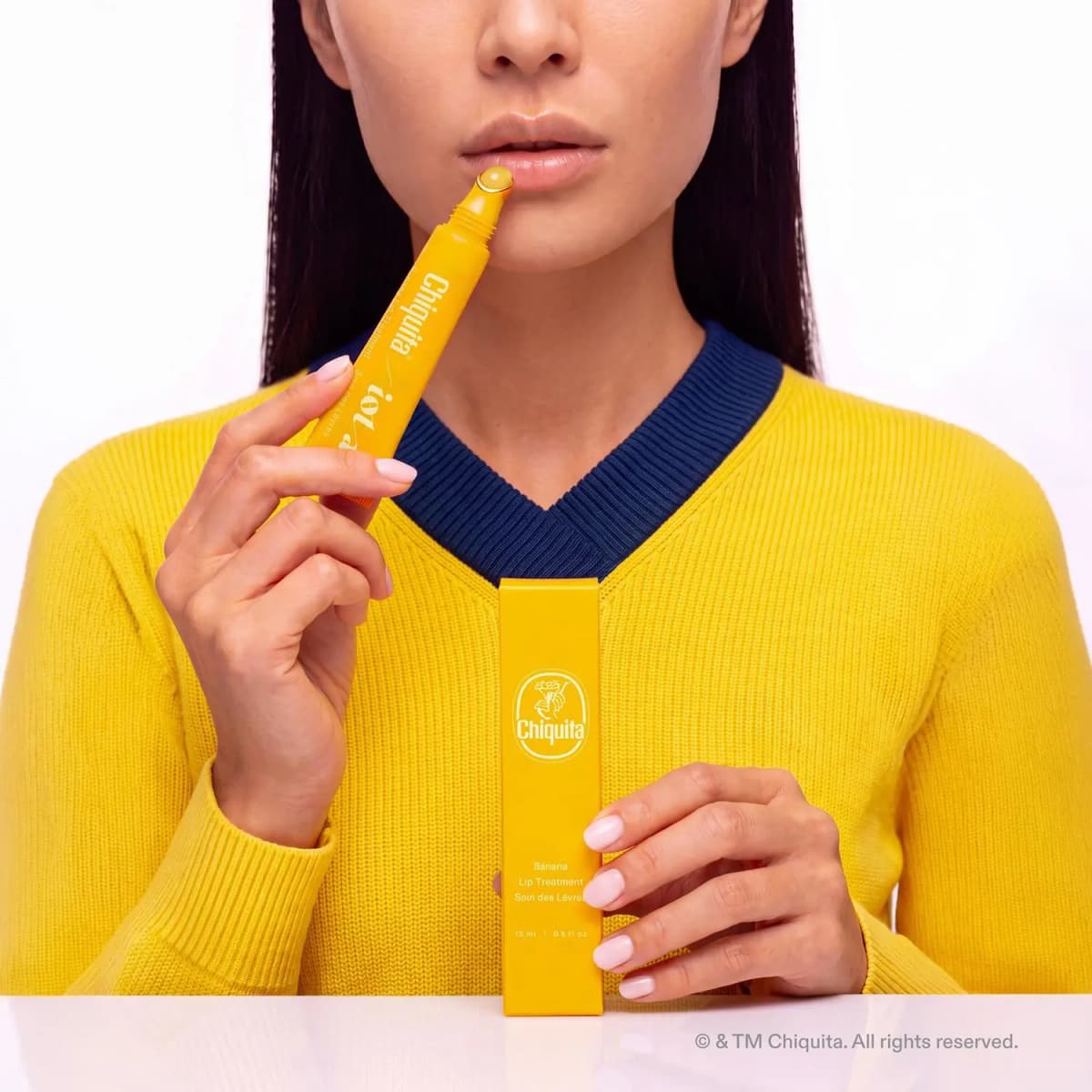 Model in a yellow sweater with navy collar applying the tube while holding the 15 ml box
