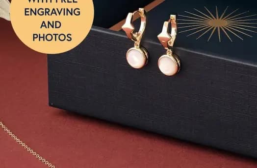 Gold drop earrings with pale pink stones resting against a navy box, with a free engraving and photos badge
