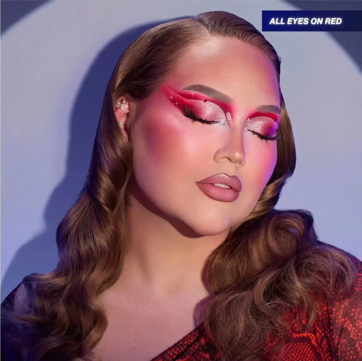 NikkieTutorials with eyes closed, showing silver glitter lids framed by red graphic wings, rhinestones and rosy blush