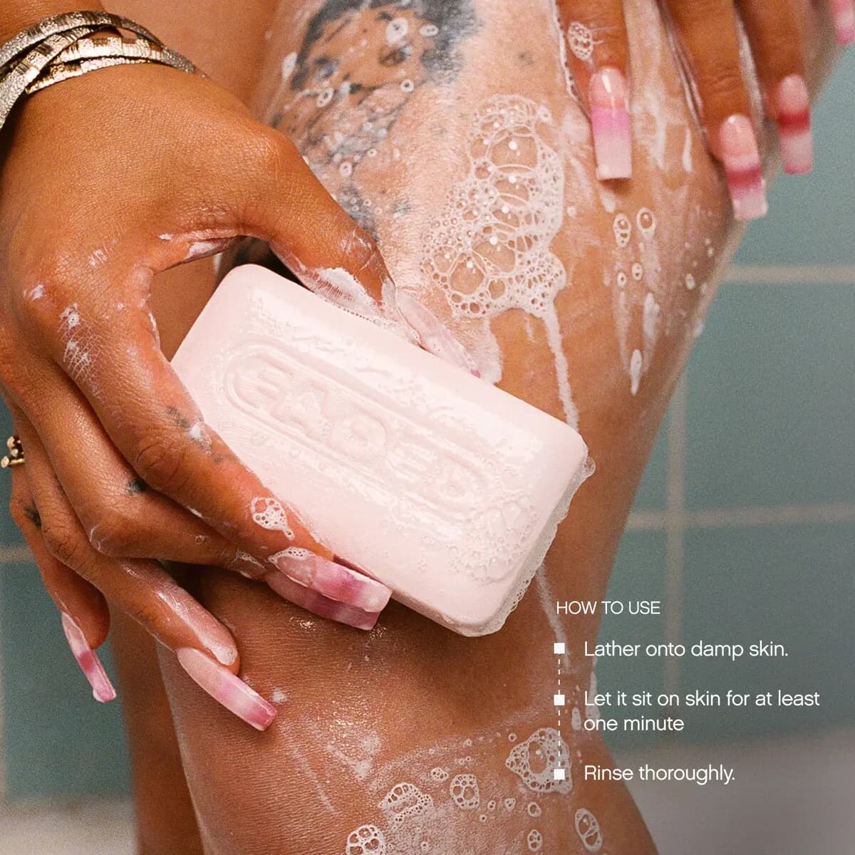 Hand lathering the pink Faded bar on a soapy leg in a tiled shower, with how-to-use steps for the bar