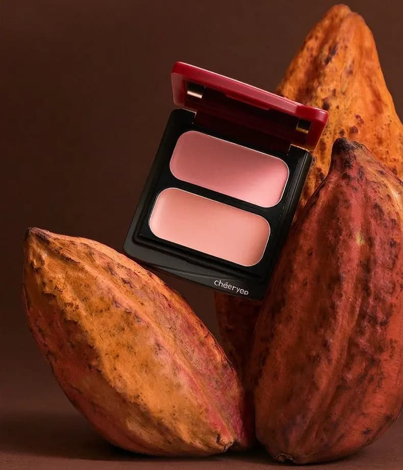 Open red-lidded chéeryep duo compact with pink and peach cream blush propped between orange cocoa pods on brown