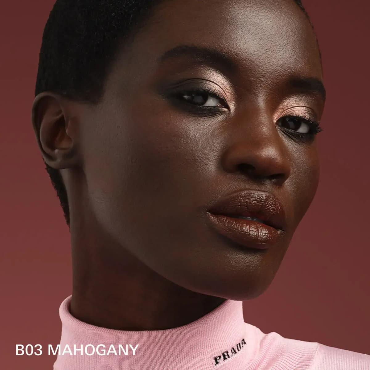 Model with deep skin wearing peachy shimmer eyes and glossy brown lips in a pink Prada turtleneck, labelled B03 Mahogany