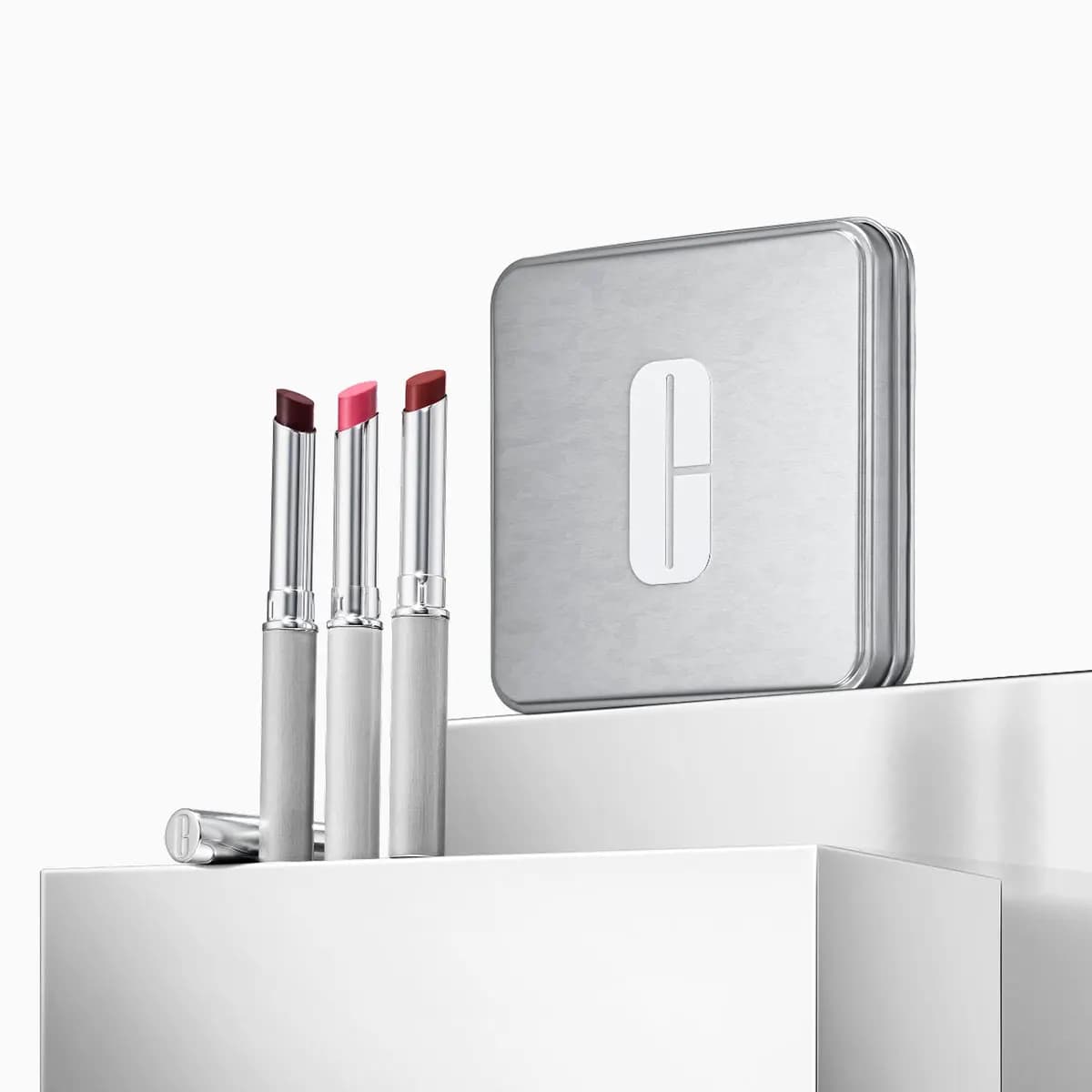 Three slim lipsticks in plum, pink and brick red beside a silver tin with the Clinique C logo