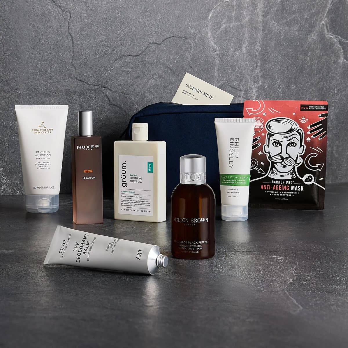 GLOSSYBOX Grooming Gift Guide products lined up on grey stone in front of a navy grooming bag with a Summer Mink card