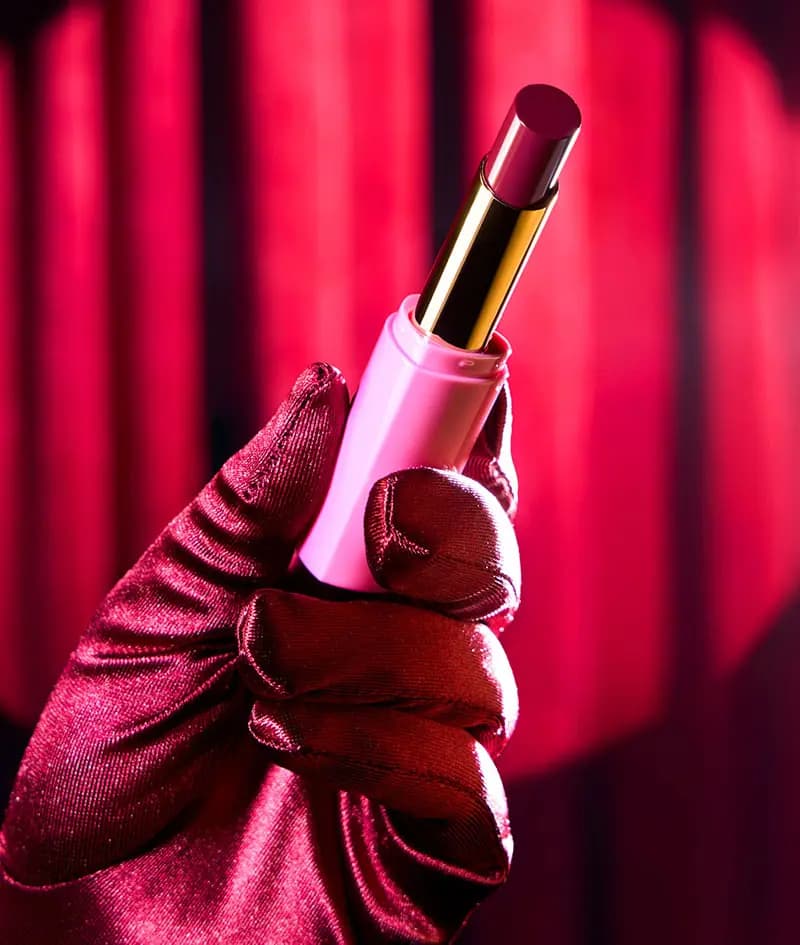 Satin-gloved hand holding the pink gloss stick with a berry bullet against glowing red curtains