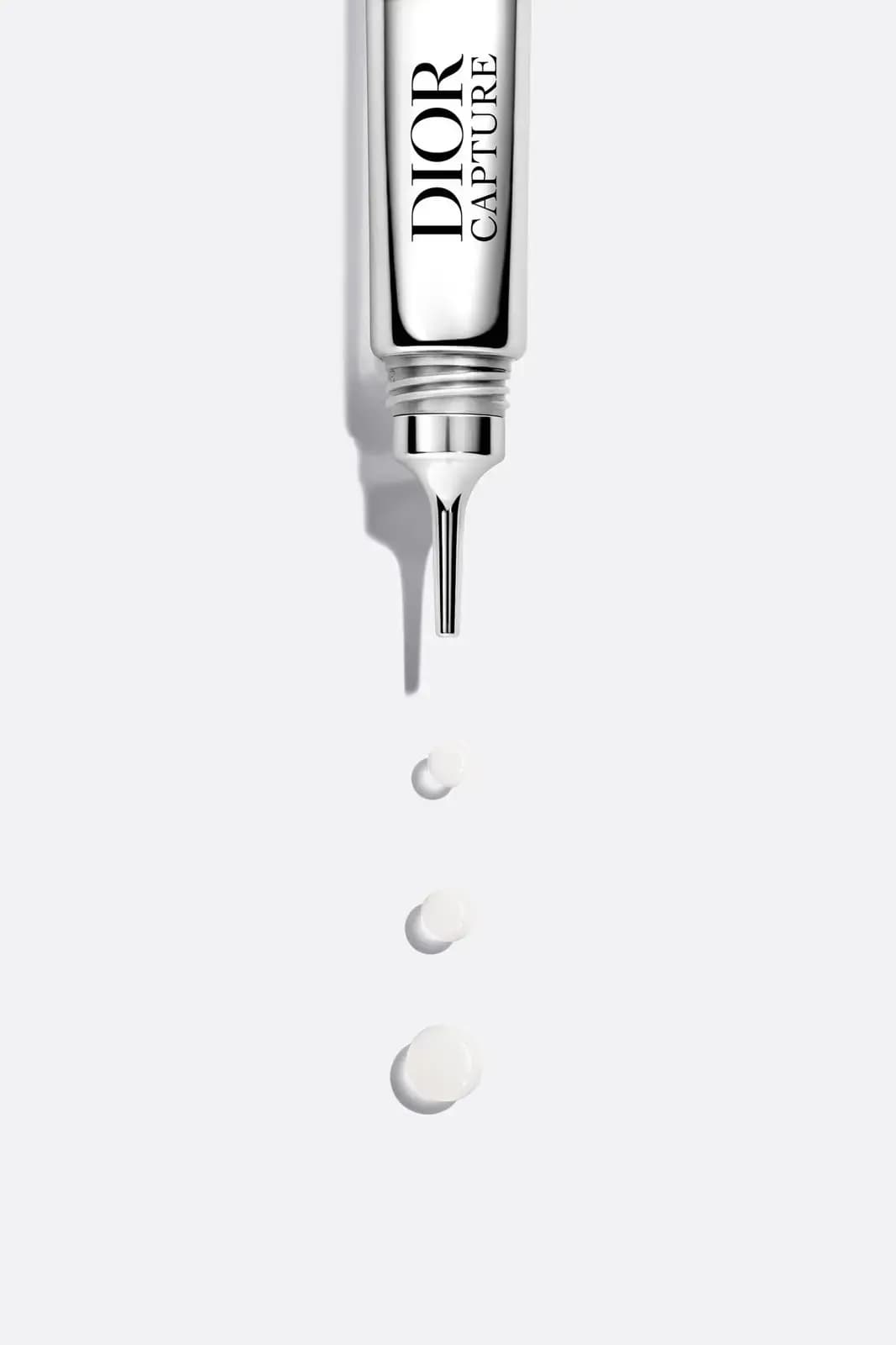 Uncapped tube held tip-down above three white dots of product on a pale grey surface