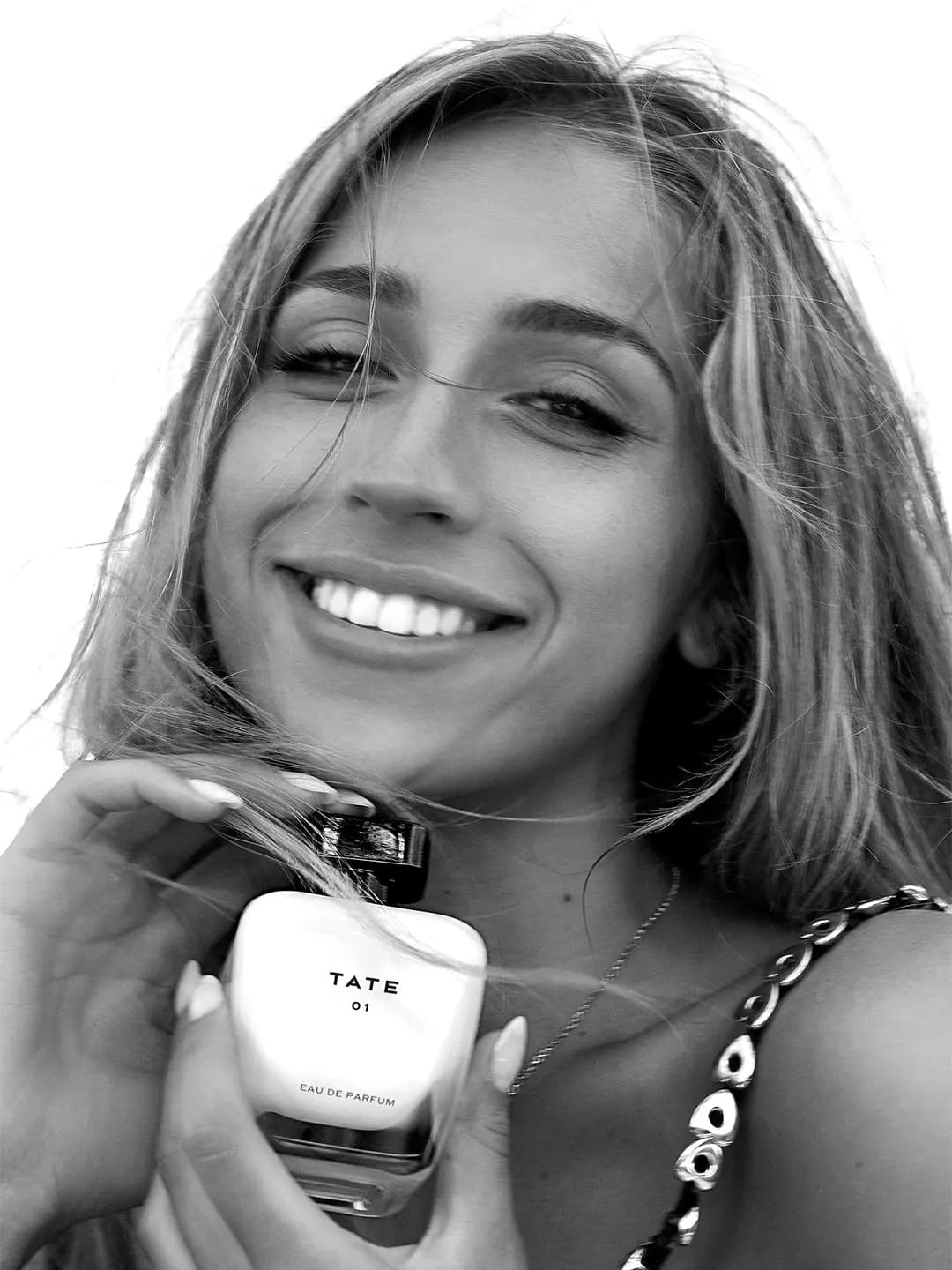 Black-and-white shot of a smiling woman with windswept hair holding a TATE 01 Eau de Parfum bottle