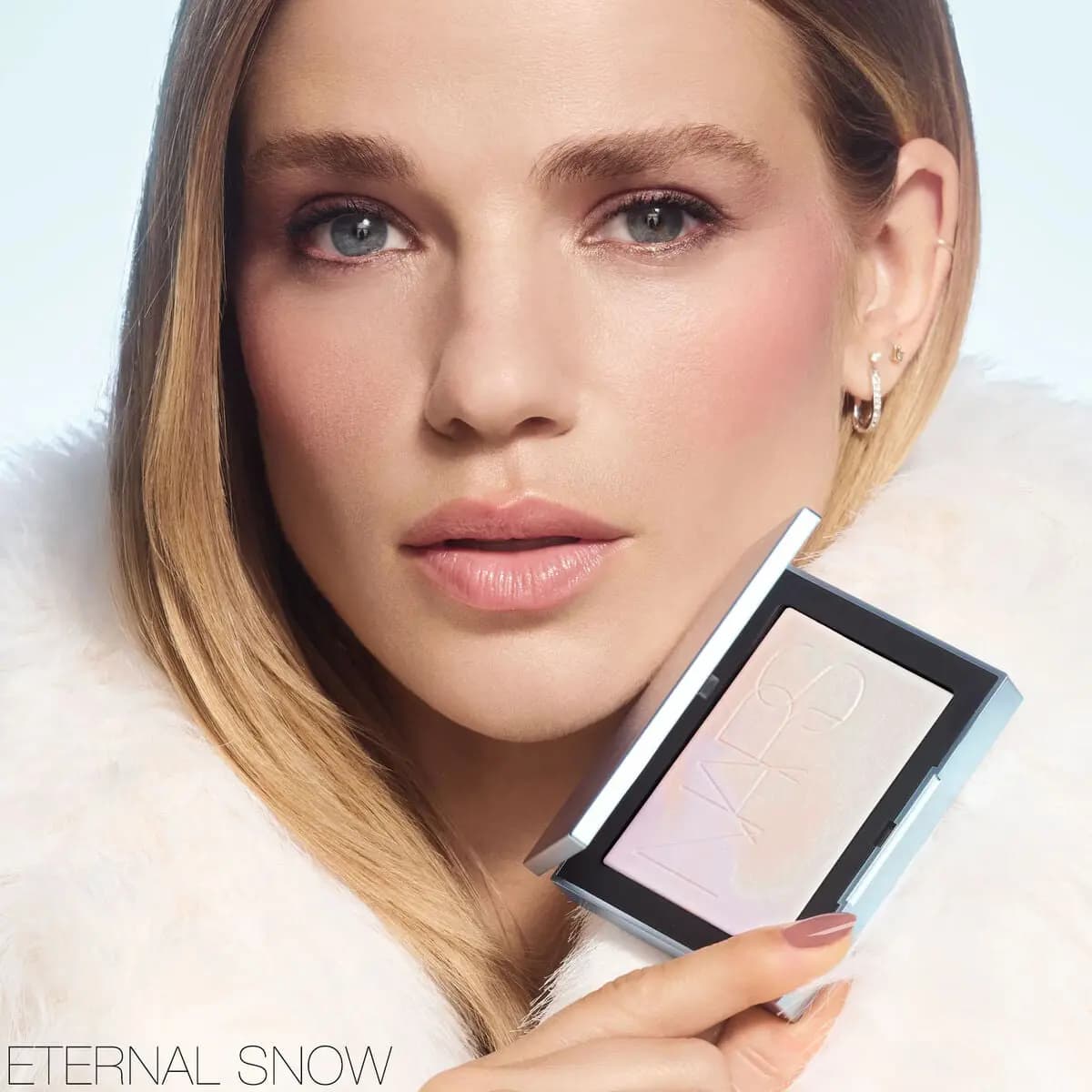 Blonde model in white fur with rosy cheeks and glowing skin holding an iridescent highlighter compact labeled Eternal Snow.