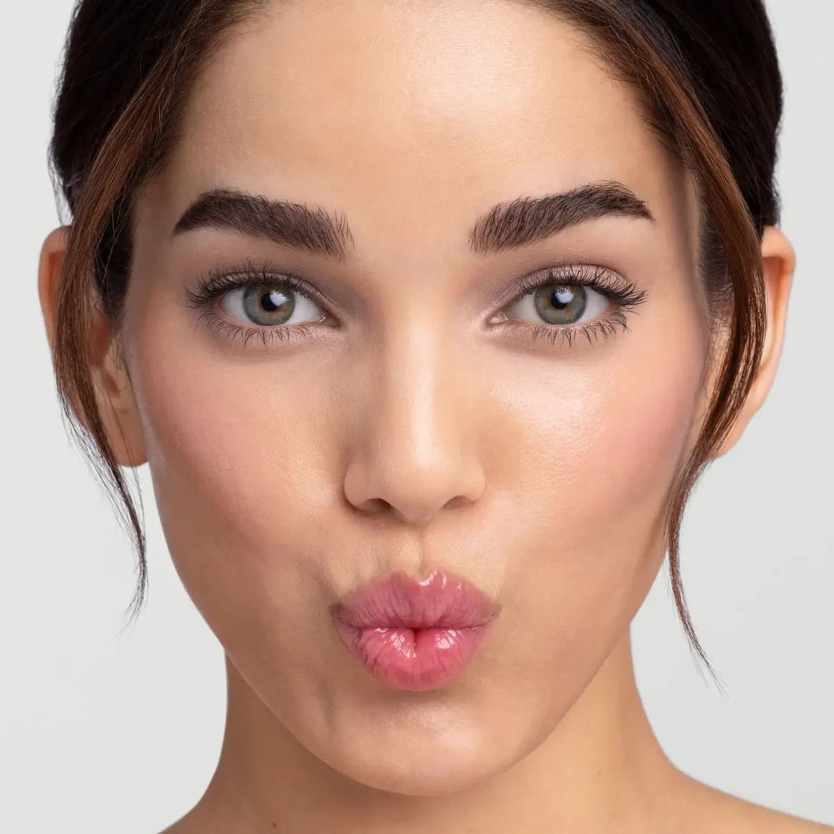 Model with dark hair pulled back and green eyes pouts glossy, sheer pink lips toward the camera.