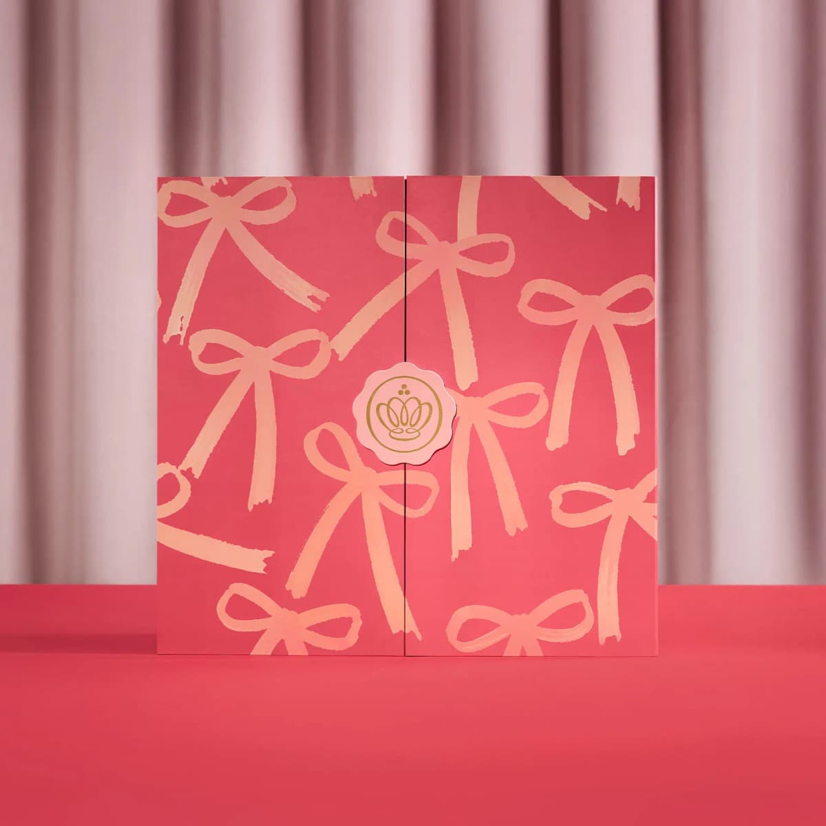 Closed pink Advent Calendar with peach bow print and gold crown seal on a red surface before a pink curtain