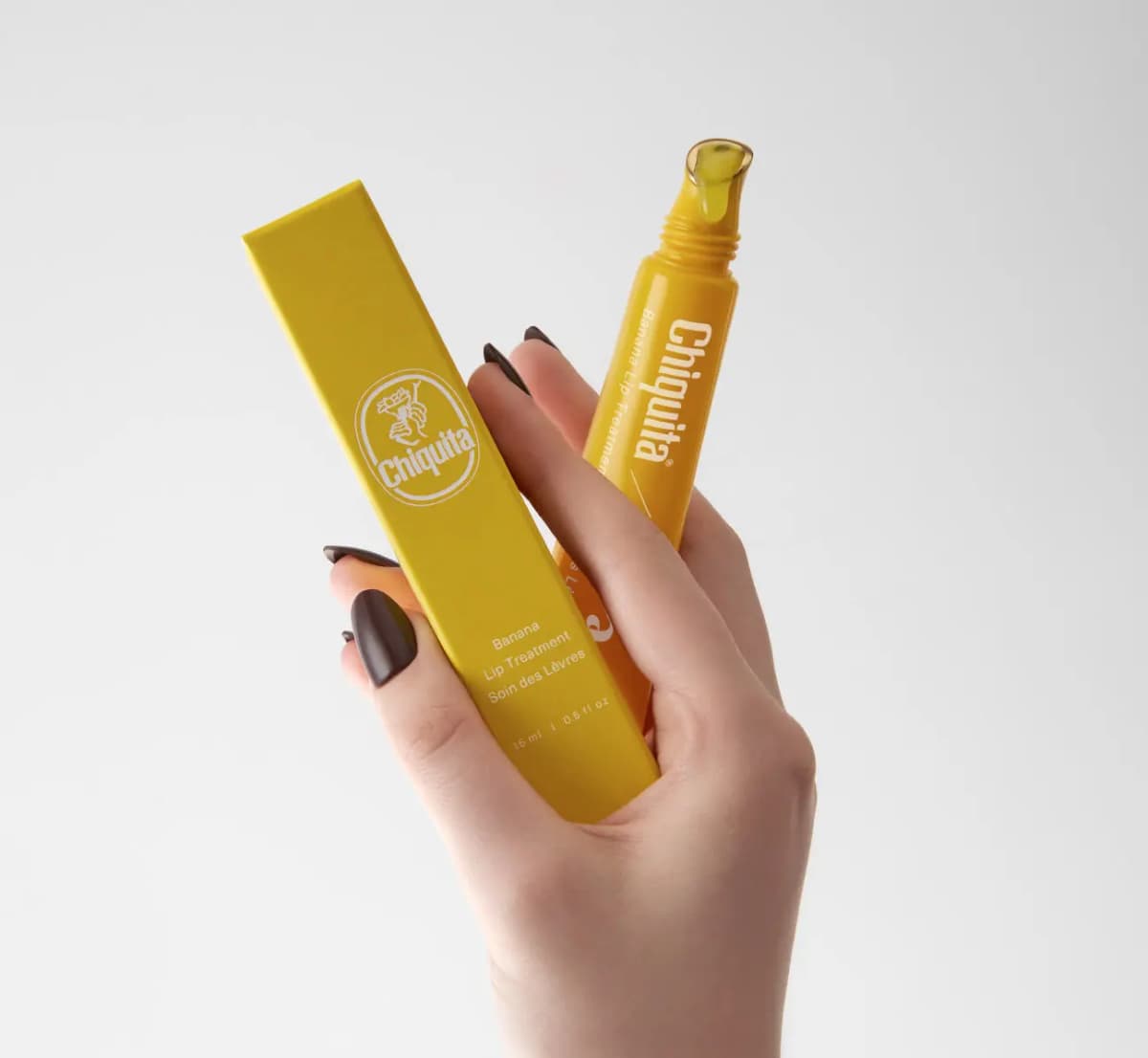Hand with dark brown nails holding the yellow box with Chiquita logo and the uncapped tube with glossy tip