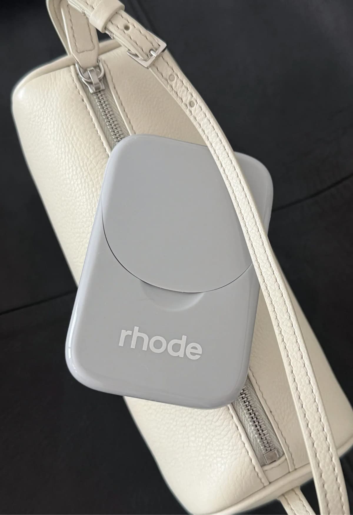 Closed pale grey case resting on a cream leather crossbody bag with silver zip against a dark background