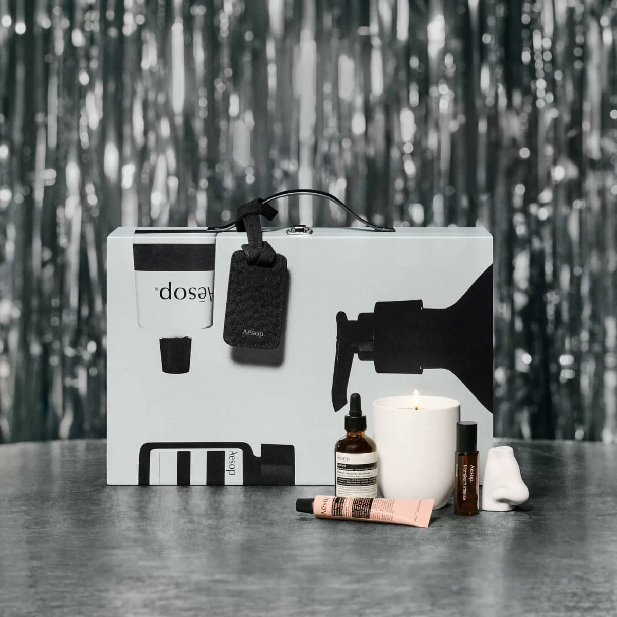 Closed white and black Aesop advent calendar suitcase with luggage tag, candle, serum, hand balm and mini fragrance in front.