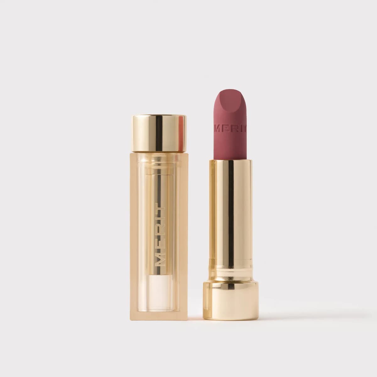 Uncapped rose-mauve MERIT matte lipstick in a gold tube beside its clear, gold-tinted cap embossed with MERIT.