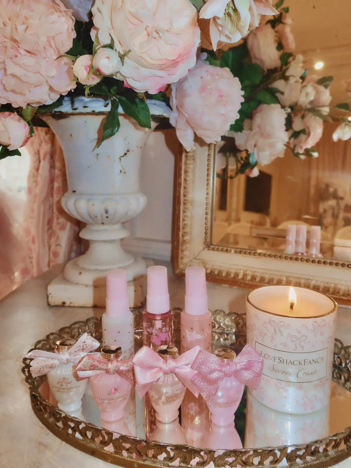 Mini perfumes with fabric bows, three pink mists and a lit Secret Crush candle on a gilded mirror tray beside pink peonies