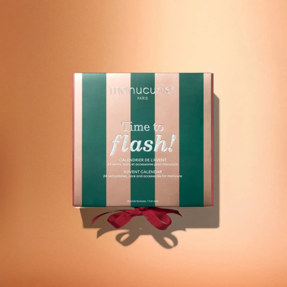 Overhead view of closed green and beige striped Time to Flash advent calendar box with red ribbon on peach background