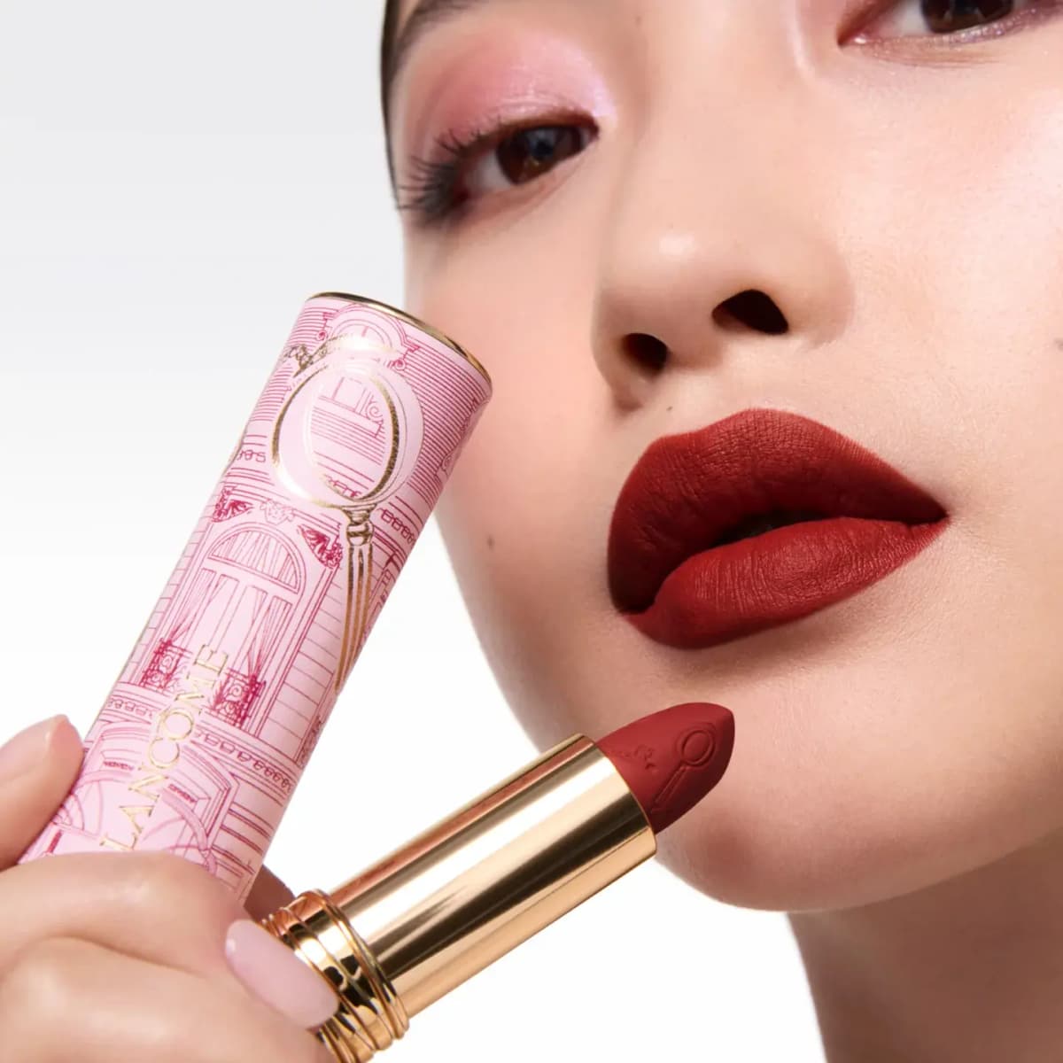 Model with pink eyeshadow and matte brick-red lips holds the lipstick and its pink Parisian facade sleeve