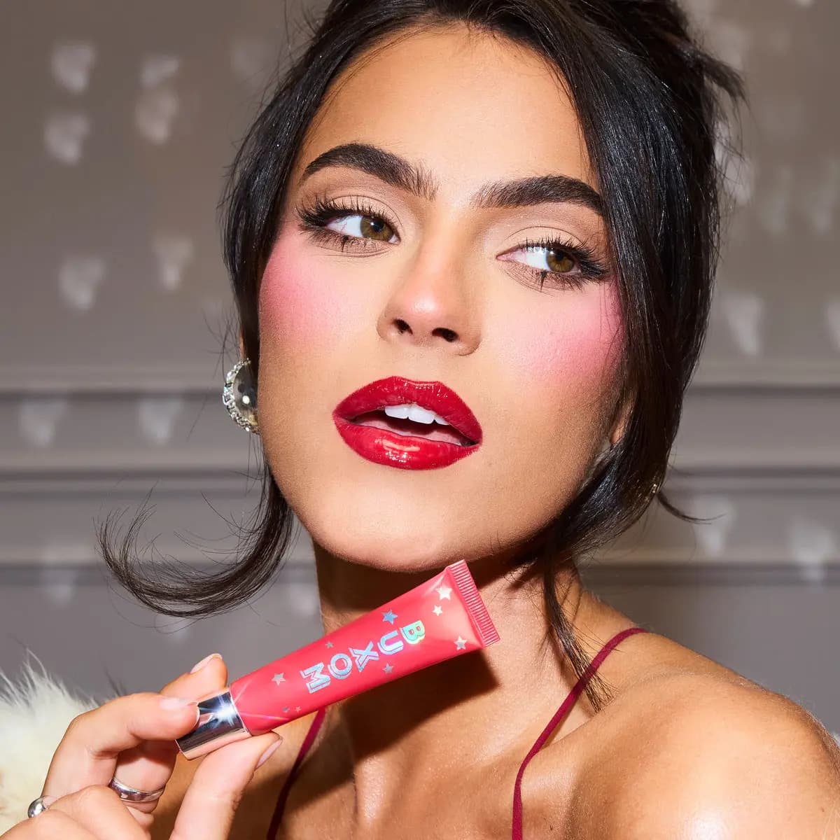 Model with dark hair and bold red glossy lips holding the pink BUXOM lip tint tube
