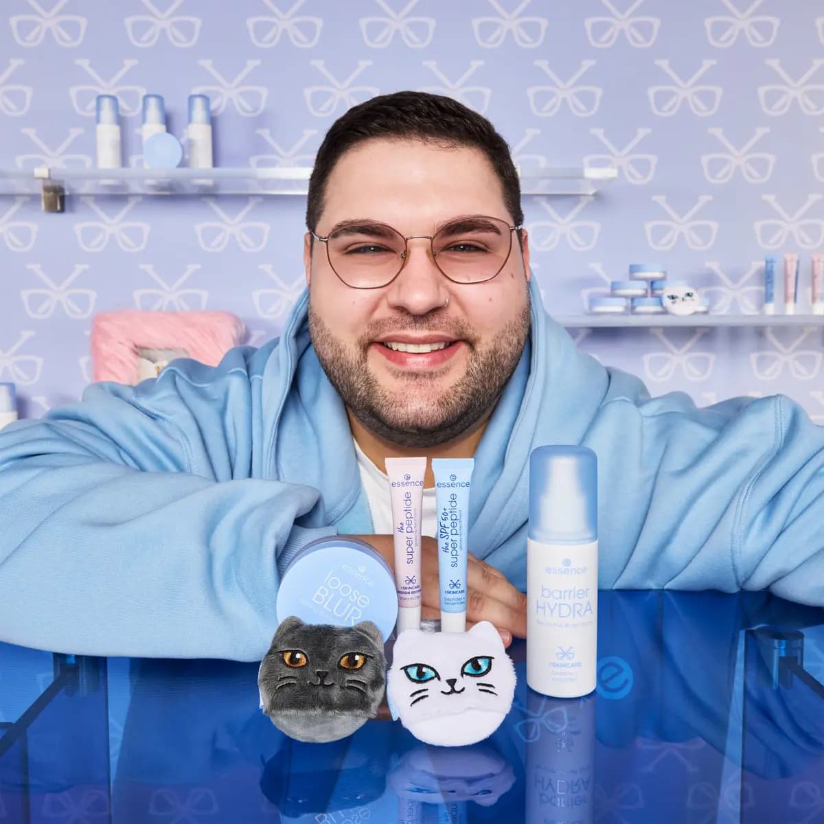 Leon xskincare in glasses and a blue hoodie, smiling behind the collection on a glossy blue table, glasses-print wall behind