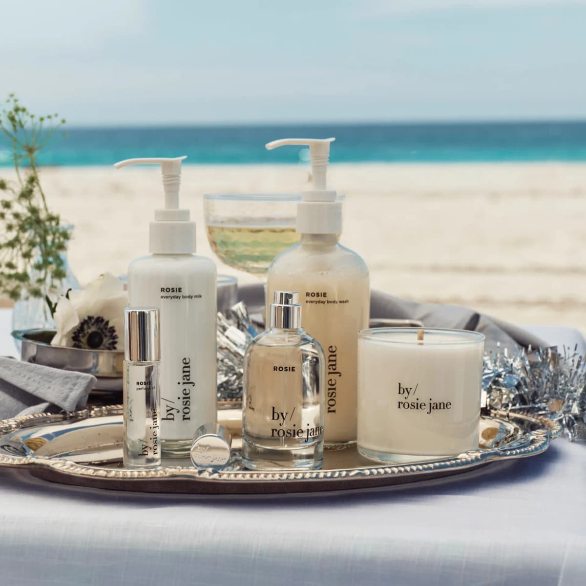 Rosie perfume oil, perfume, body milk, body wash and candle on a silver tray at a beach table with sea behind
