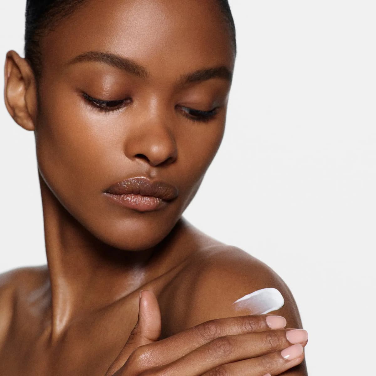 Close-up of a woman with sleek hair and glossy lips applying white lotion to her bare shoulder