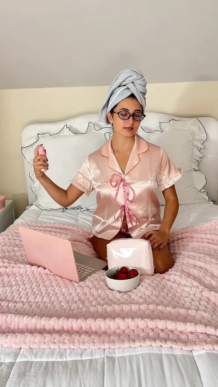 Woman in pink satin pajamas and hair towel sitting on a bed with a pink laptop, strawberries, and cosmetic pouch