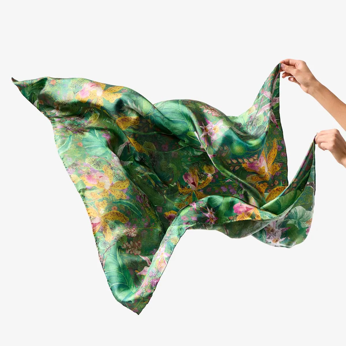 Hands holding up a satin green scarf printed with pink and yellow orchids and tropical leaves on a white backdrop
