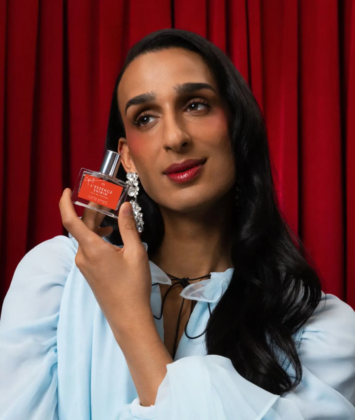 Cyrus Veyssi in a pale blue blouse and crystal earrings holds a L'Essence Shirin bottle near their cheek against red drapes