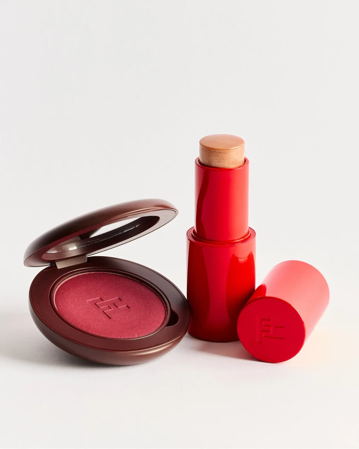 Open brown compact with deep berry pressed powder beside an uncapped red stick of golden balm and its FH-embossed cap