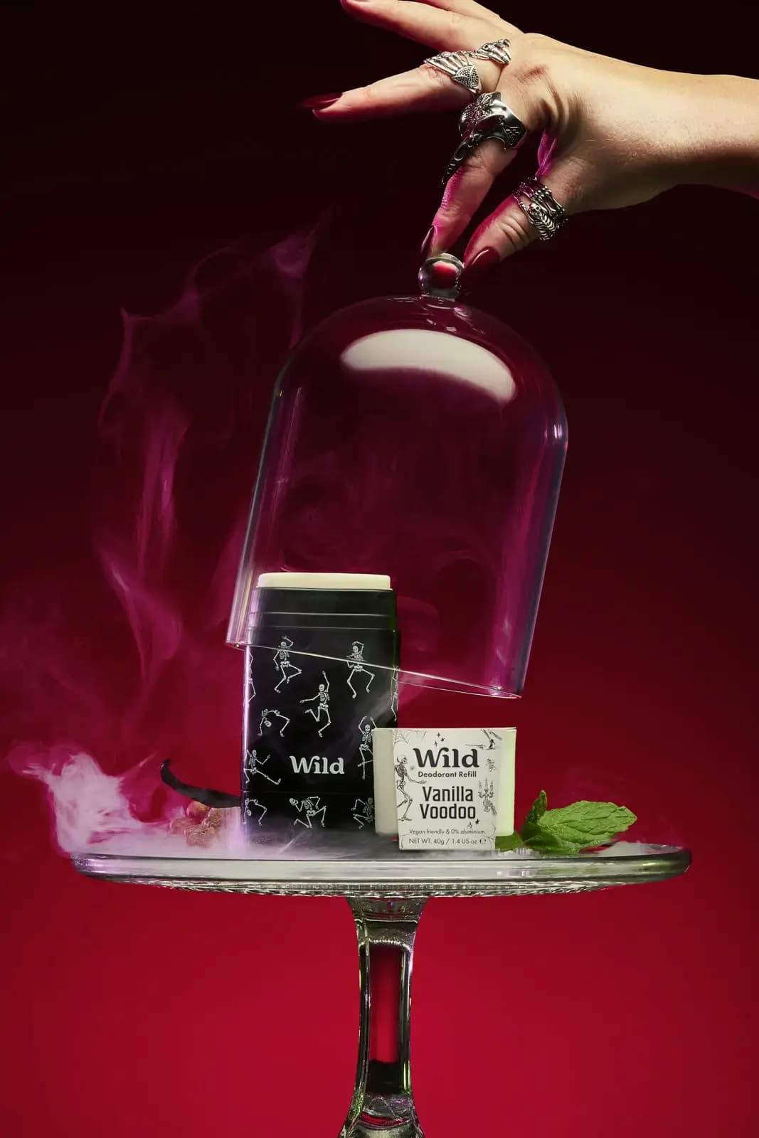 Ringed hand lifts a glass cloche off a Wild Dancing Skeletons stick case and Vanilla Voodoo refill on a smoky cake stand