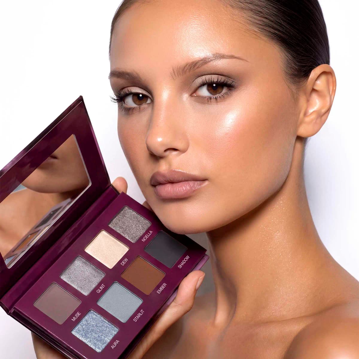 Model with glowing skin and soft neutral eyes holding the open eight-shade Serenade palette with its mirror.