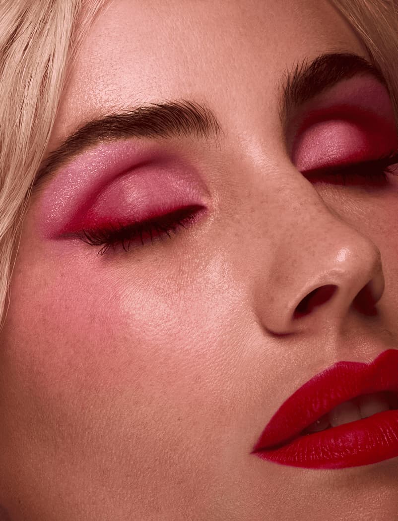 Close-up of closed eyes with vivid pink shimmer eyeshadow and bold red matte lips on fair freckled skin.