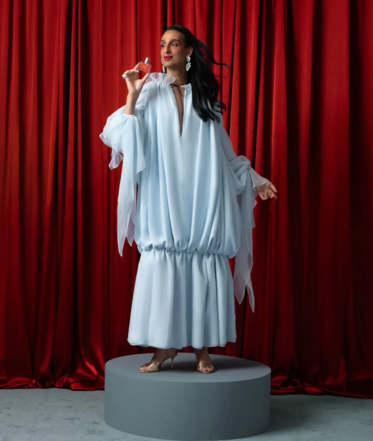 Cyrus Veyssi in a voluminous pale blue chiffon gown and gold heels poses on a grey plinth with the perfume, red curtain behind