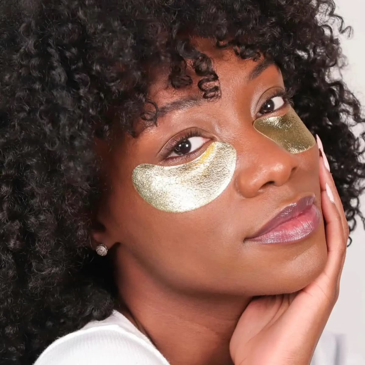Smiling model with curly dark hair wearing metallic gold under-eye masks, resting a cheek on one hand