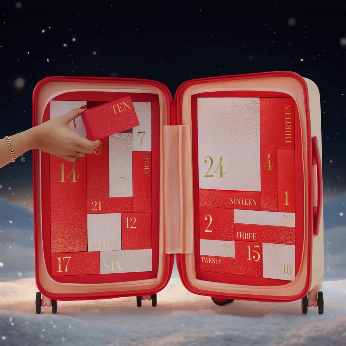 Hand lifting the red TEN box from the open suitcase, revealing gold-numbered red and pale pink drawers against snow