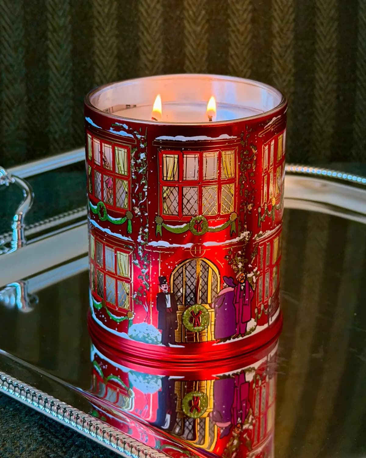 Lit red townhouse candle tin with two flames reflected on a silver mirrored tray against dark green fabric
