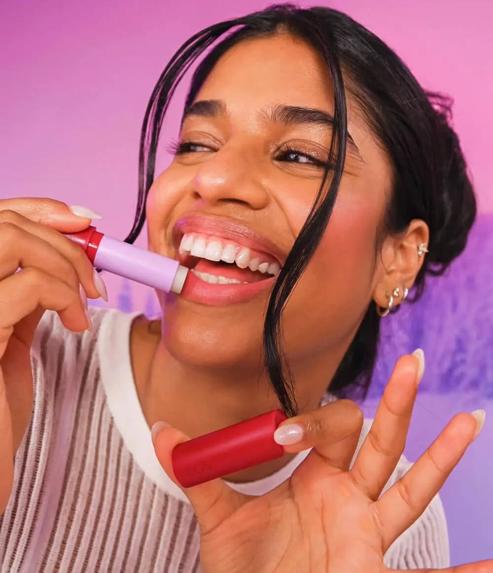 Smiling woman applying a lavender lip balm, holding a red eos cap, against a pink and purple backdrop