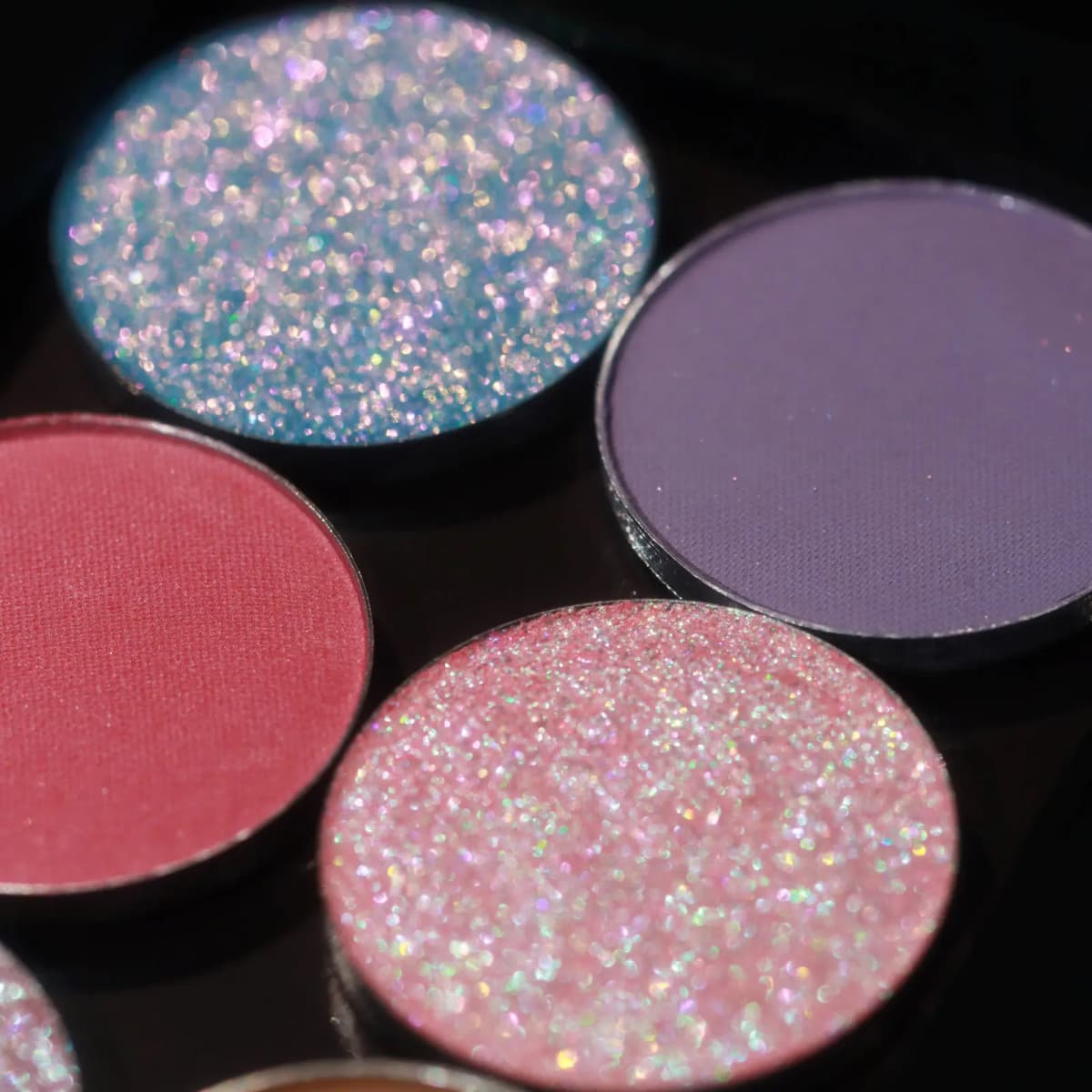 Close-up of four eyeshadow pans in blue-pink glitter, matte purple, matte berry, and pink holographic sparkle