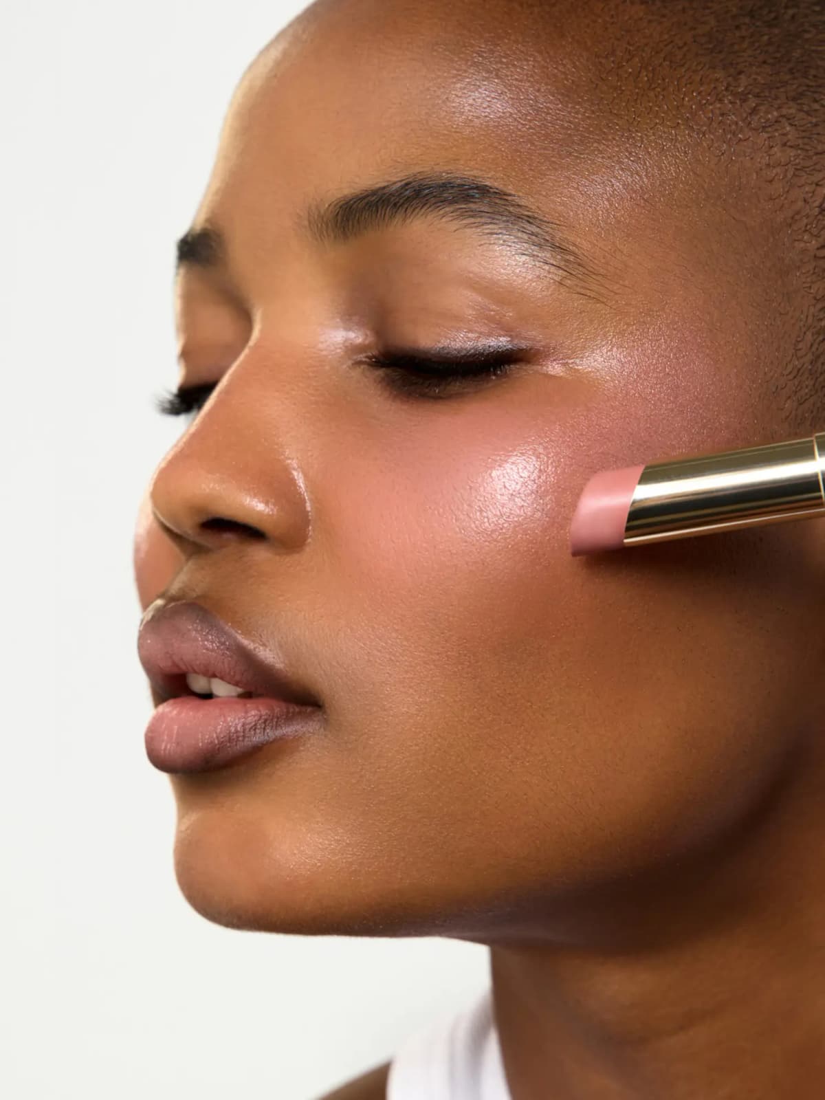 Pale pink balm stick in a gold tube swept onto a model's cheek, leaving a dewy pink glow