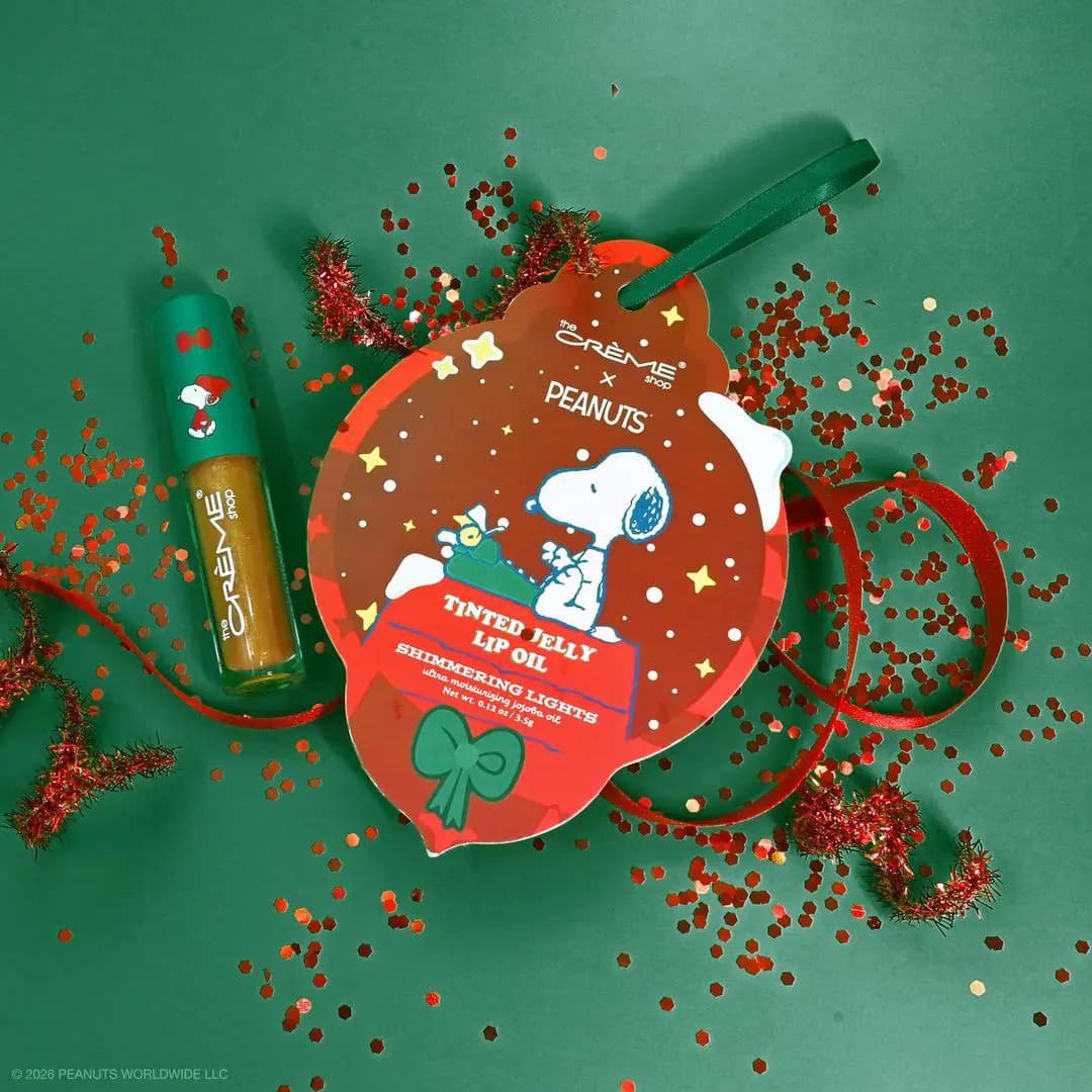 Shimmering Lights lip oil beside its ornament card on green with red glitter, tinsel and ribbon.