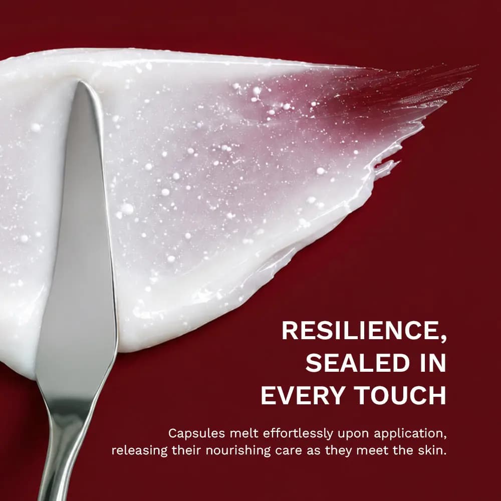 Silver spatula spreading white cream flecked with melting capsules across a deep red background