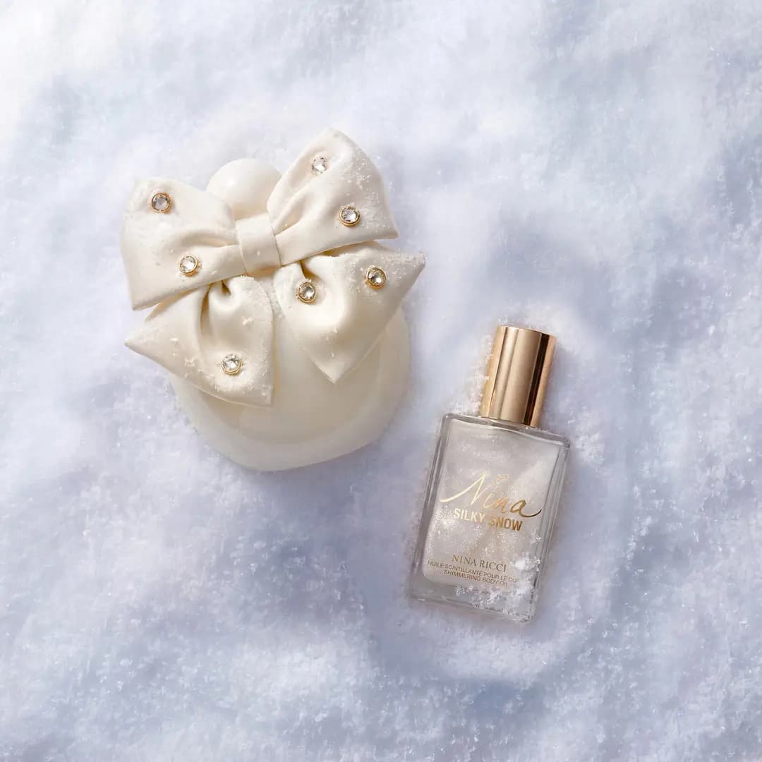 Overhead view of the bow bottle beside a gold-capped Silky Snow Shimmering Body Oil, both resting in powdery snow