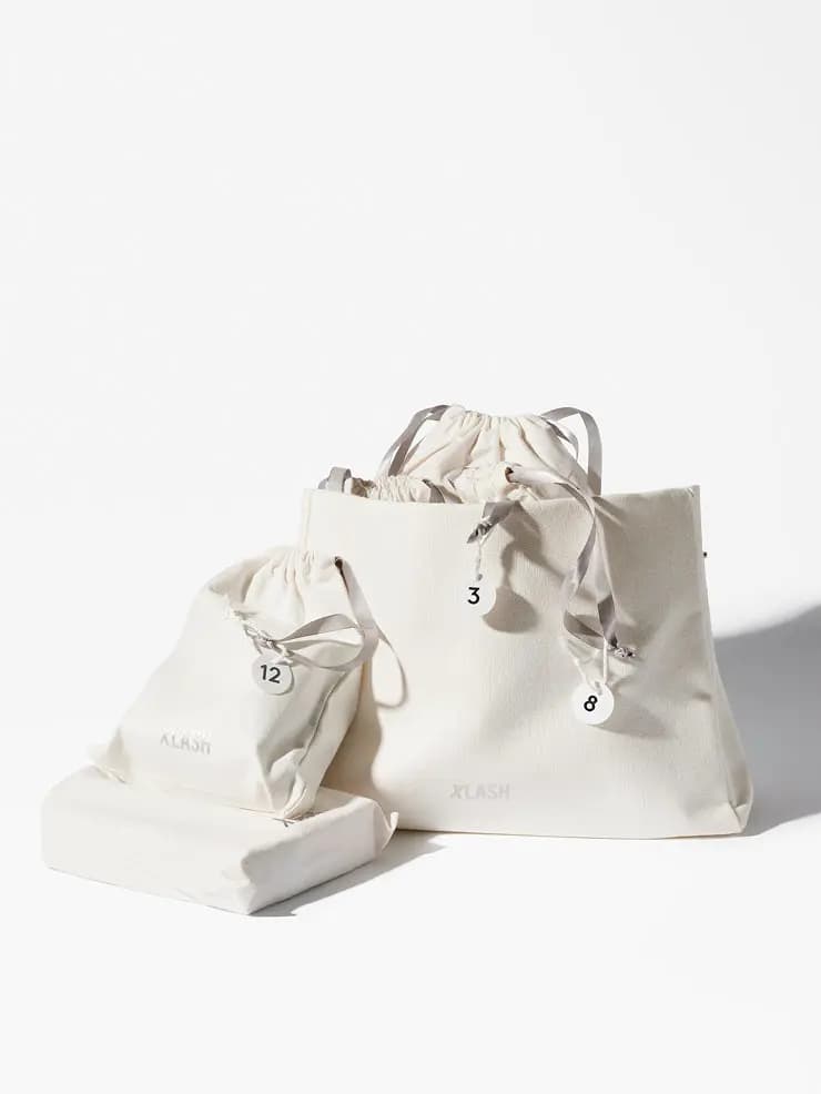 Cream Xlash beauty bag holding numbered drawstring pouches with grey satin ribbons, a 12 pouch and wrapped box beside it