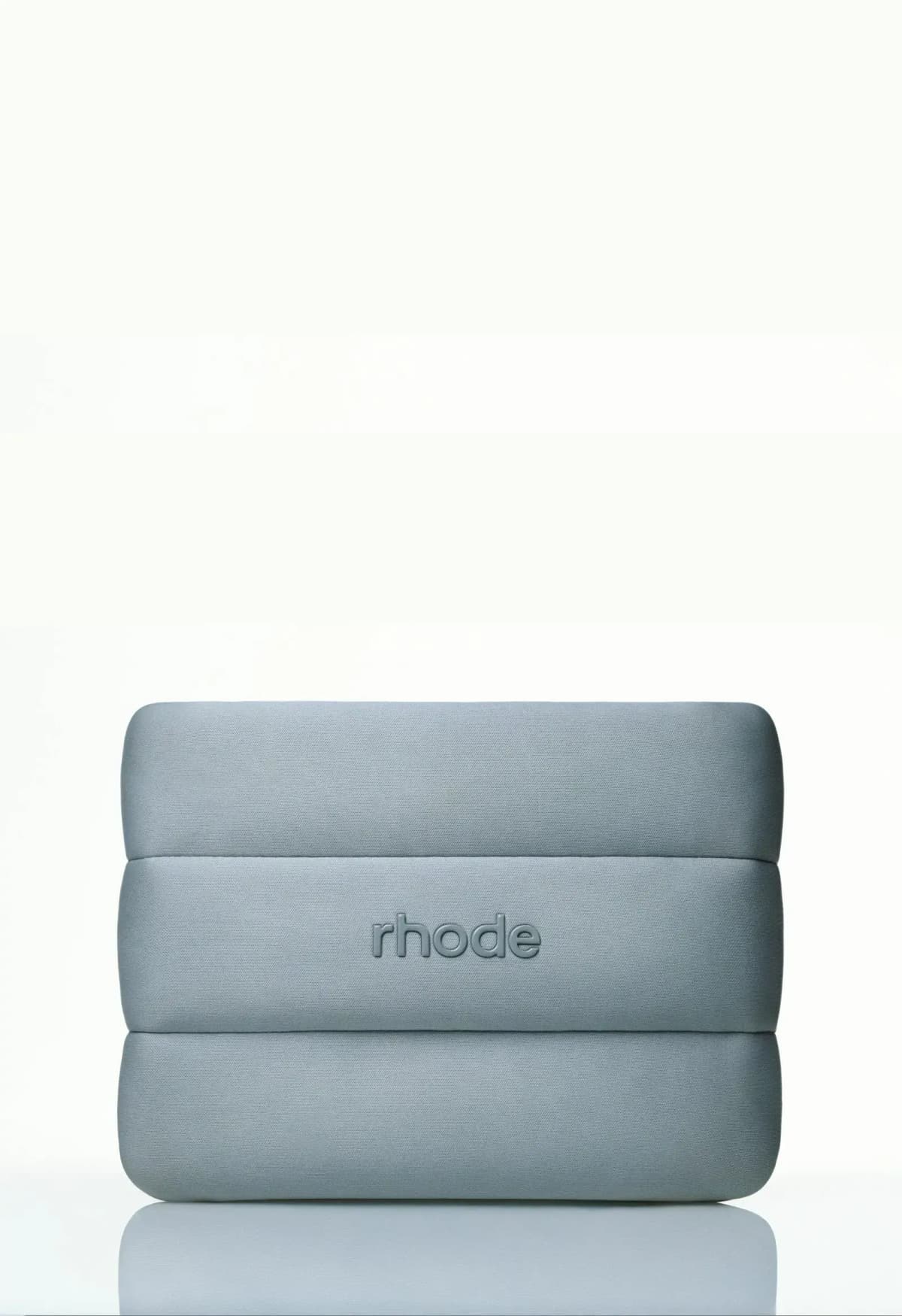 Blue-grey puffy pouch with embossed rhode logo standing upright on a reflective white surface