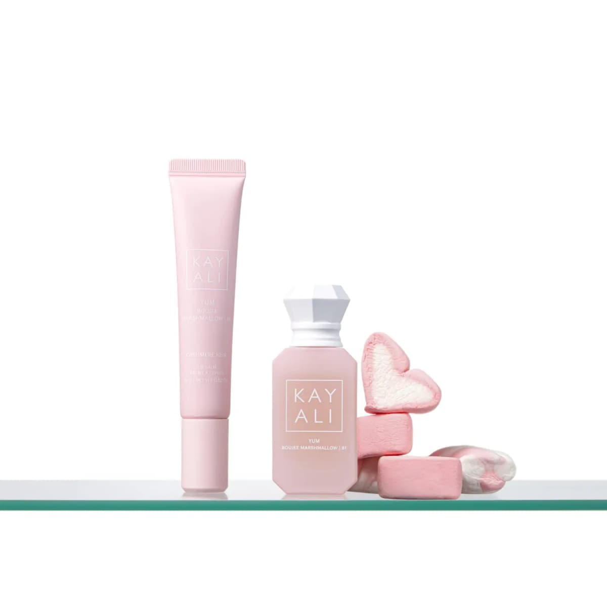 Pink Cashmere Kiss lip balm and Boujee Marshmallow perfume on a glass shelf next to pink and white marshmallows
