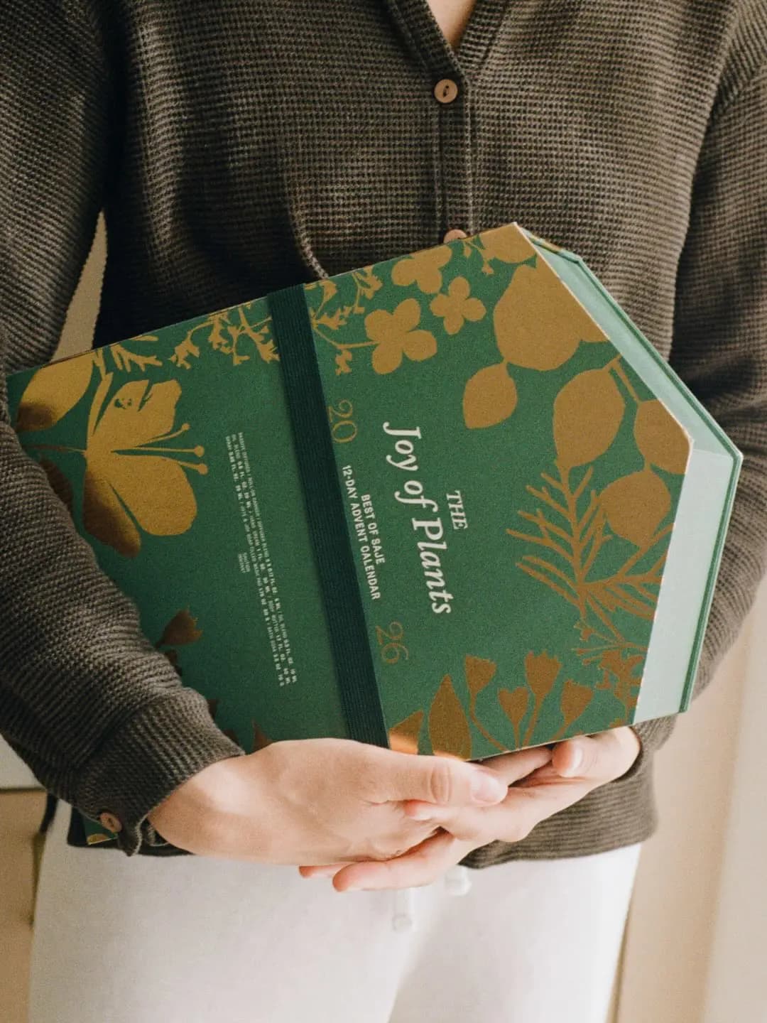 Person in a brown knit cardigan holding the green and gold foil Saje Joy of Plants advent calendar box with an elastic band