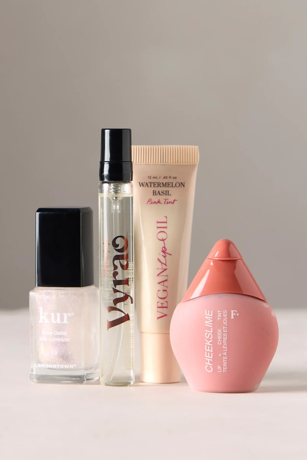 Close-up row of shimmery Kur nail luminizer, clear Vyrao spray vial, Vegan Lip Oil tube and pink Cheekslime bottle